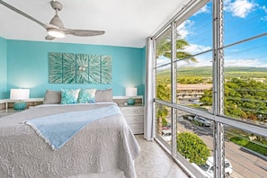 A bedroom with a large bed, a ceiling fan, a view of the city through a large window, and a tropical-themed wall art piece.