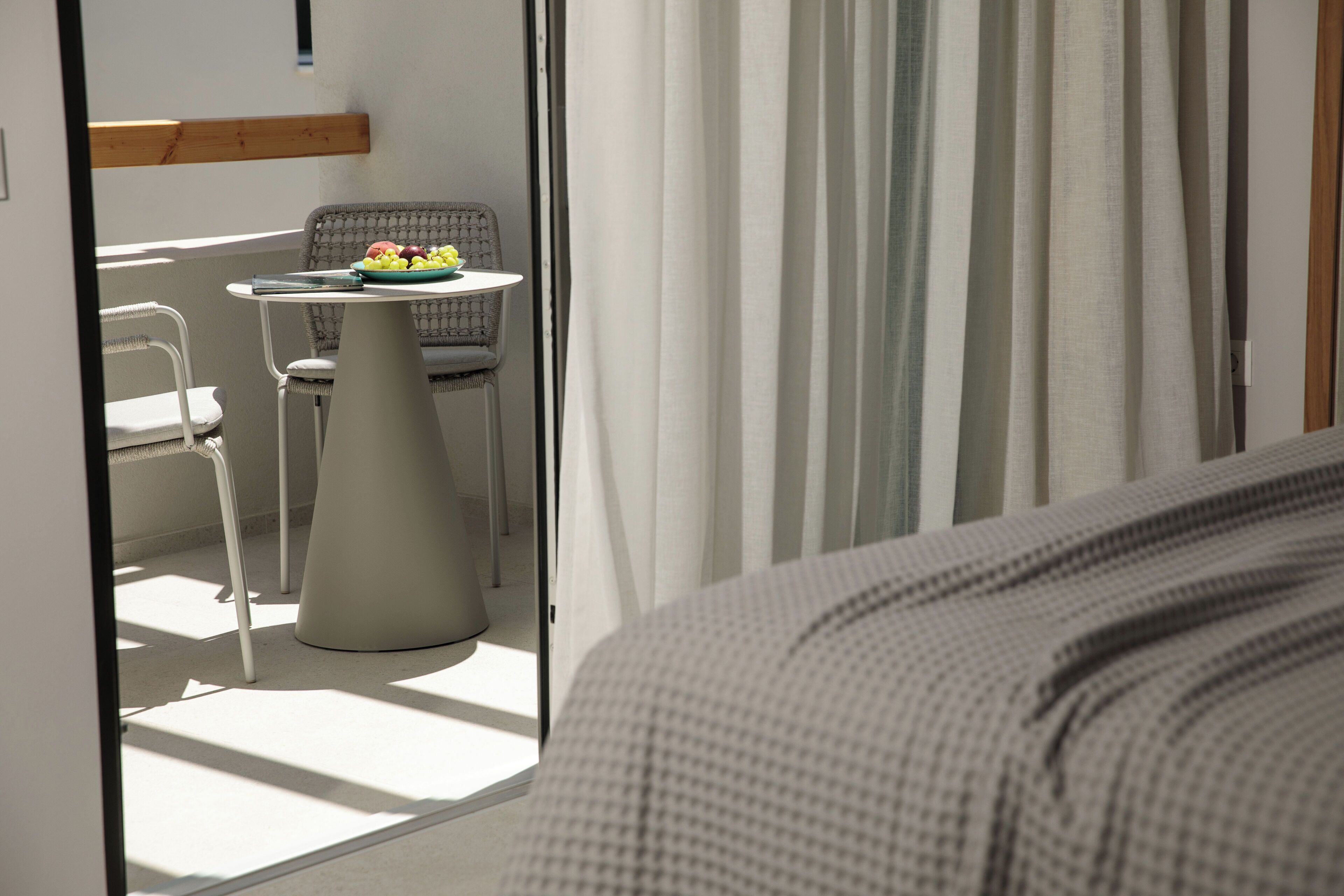 A hotel room with a bed, a small table with a fruit bowl, a chair, and a view of the outside through a window with curtains.