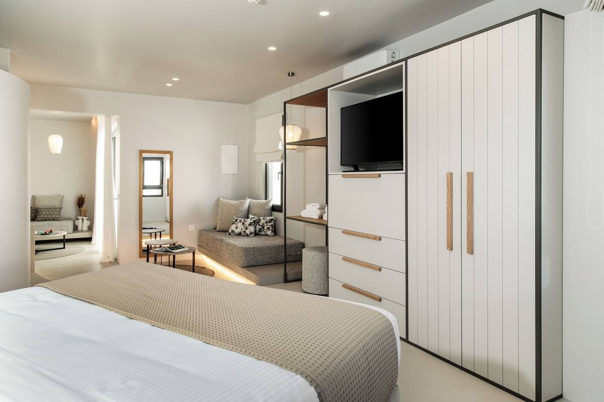 Un dormitorio moderno con una cama grande, un armario con televisión, una zona de estar con sofá y cojines, y una mesita de noche con lámpara.