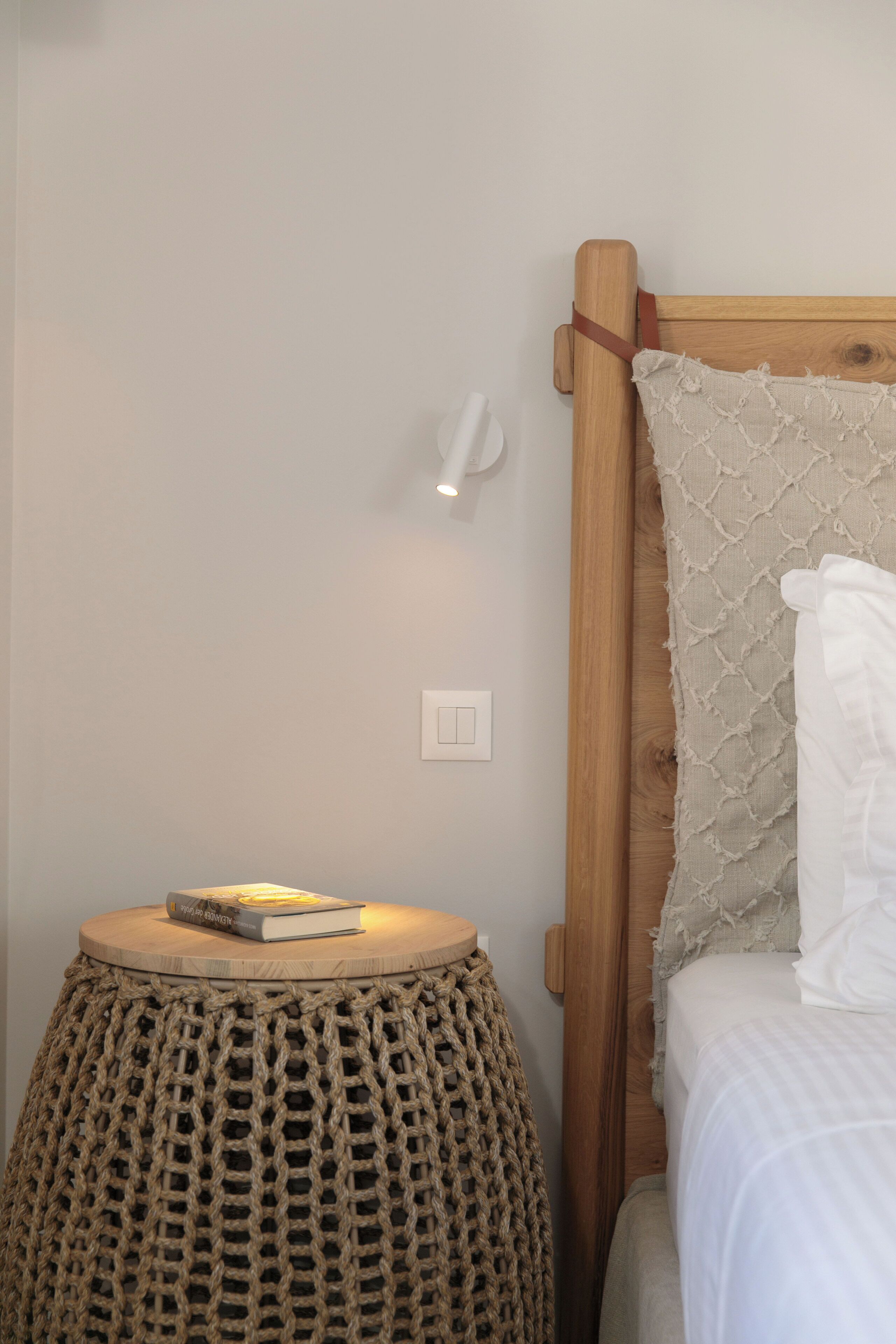 A neatly made bed with a wooden headboard, a small round table with a book, and a wall-mounted light.