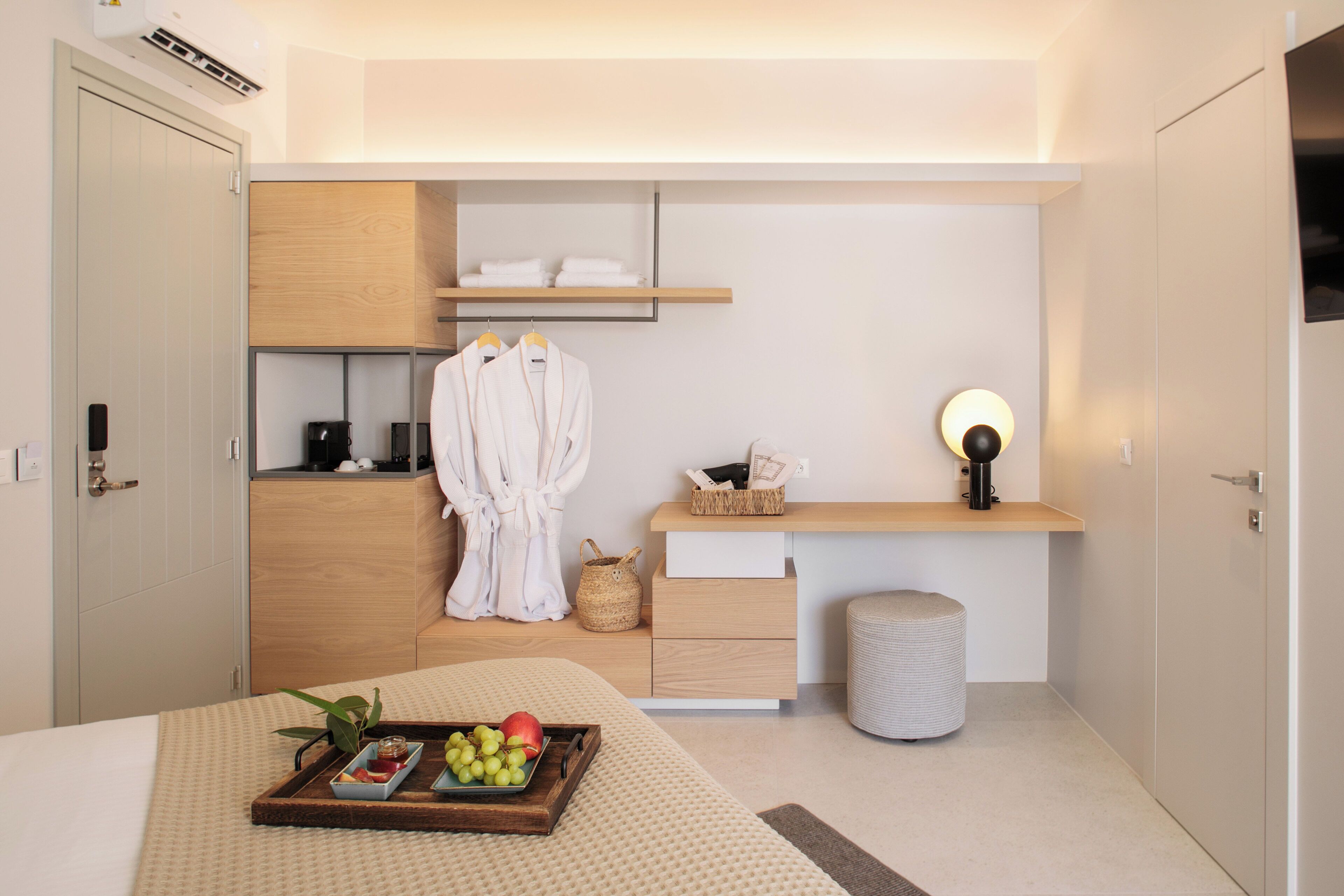 A modern hotel room with a bed, a bedside table with a tray of fruit, a robe, a shelf with towels, a waste bin, and a wall-mounted lamp.