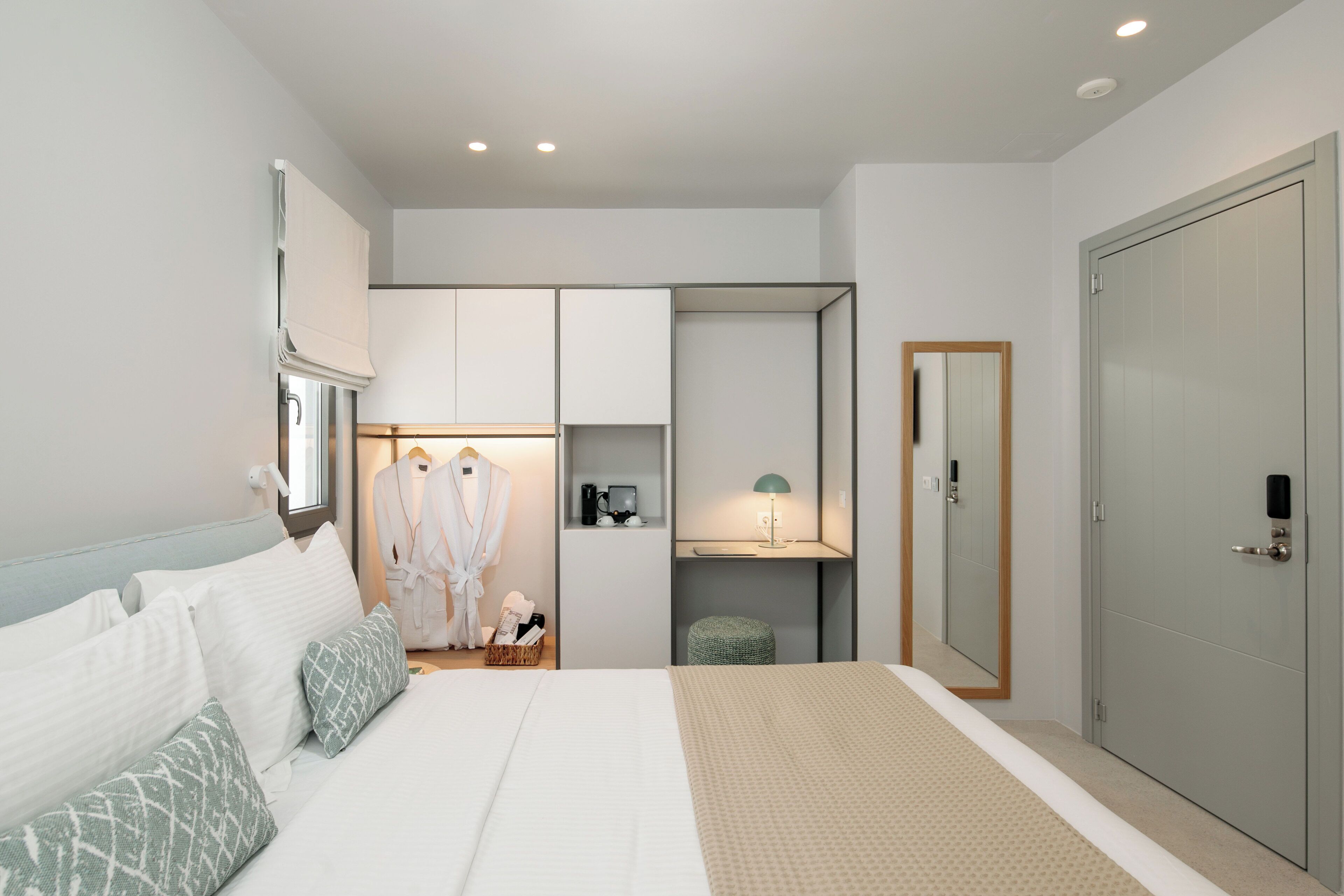 A modern hotel room with a large bed, a wardrobe, a vanity with a mirror, and a small stool.