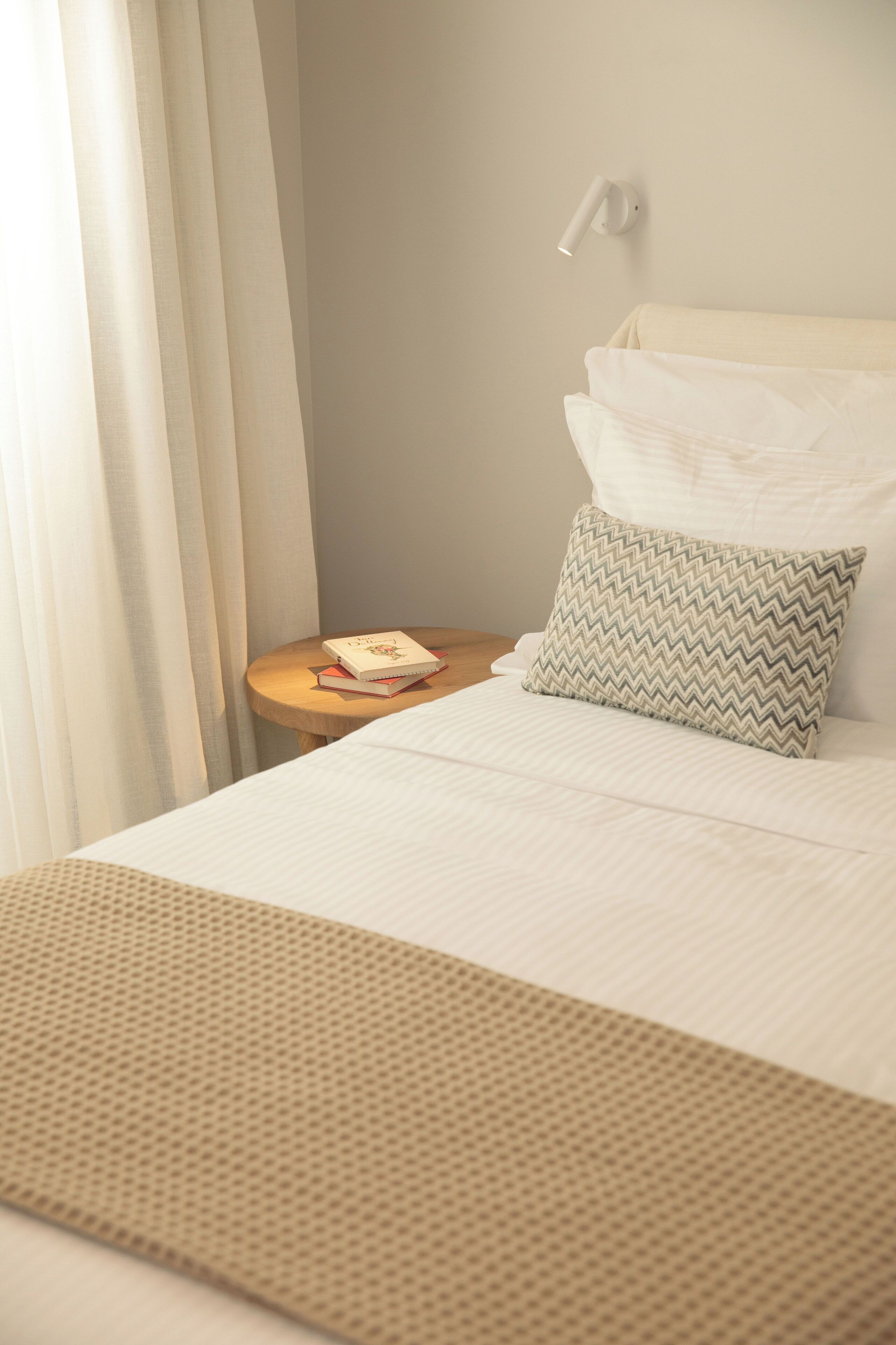 Una cama bien hecha con un cojín, una mesita de noche con un libro, y una lámpara de pared.