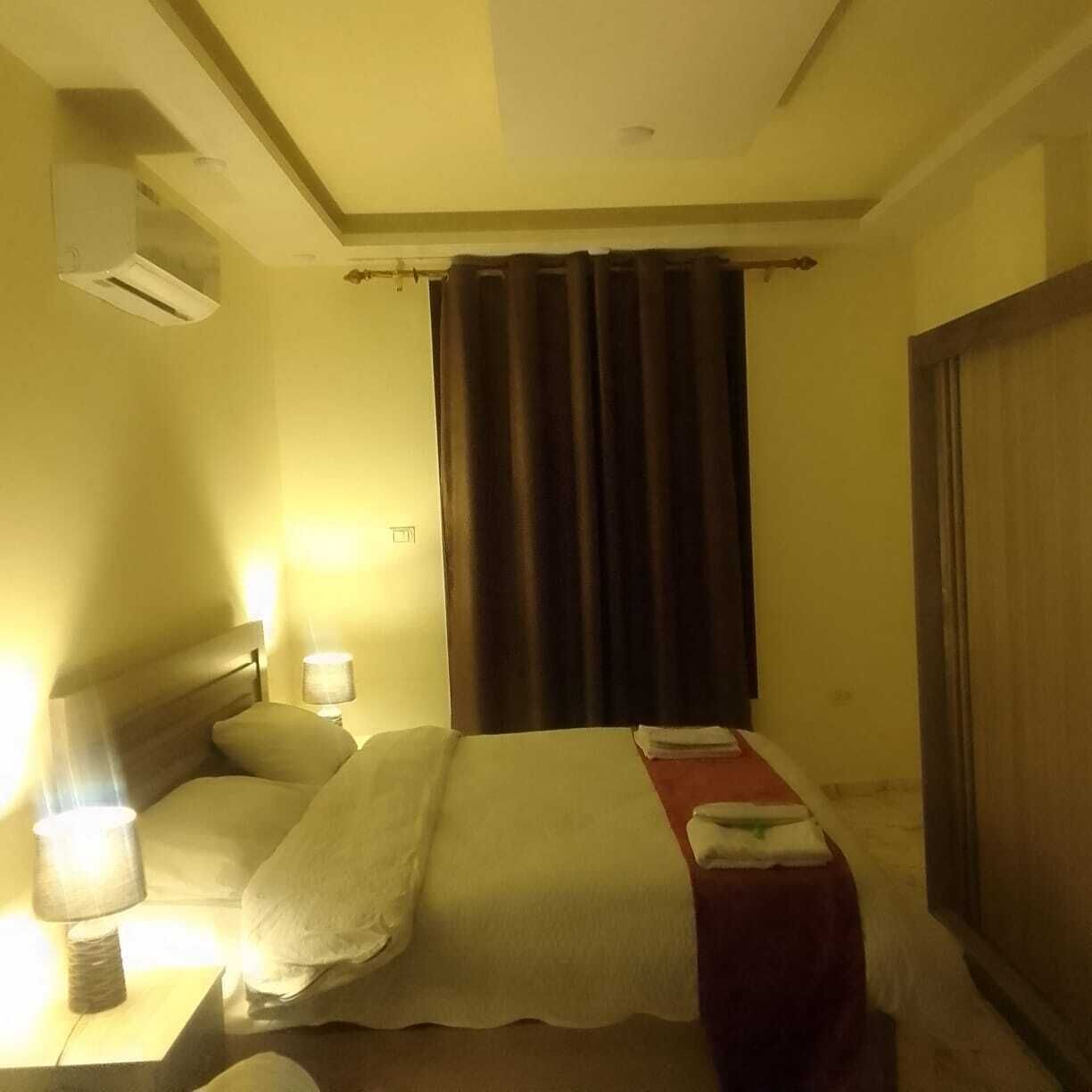 Una habitación de hotel con una cama, una mesita de noche con lámpara, una ventana con cortinas y un equipo de aire acondicionado.