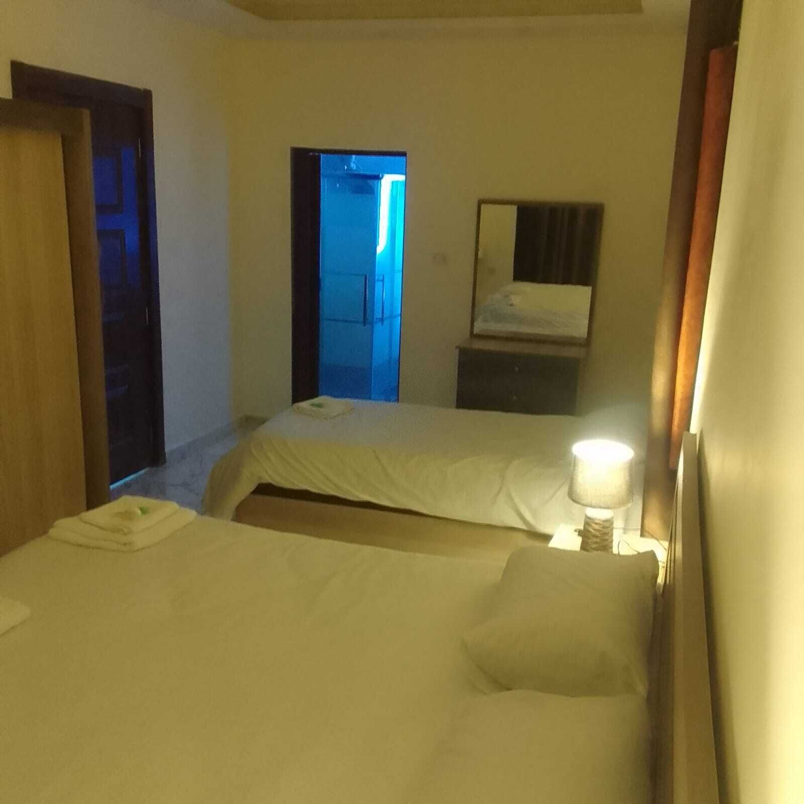 A hotel room with two beds, a bedside table with a lamp, a mirror, and a door leading to another room.