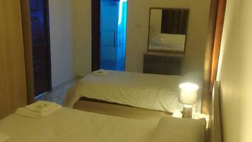 A hotel room with two beds, a bedside table with a lamp, a mirror, and a door leading to another room.