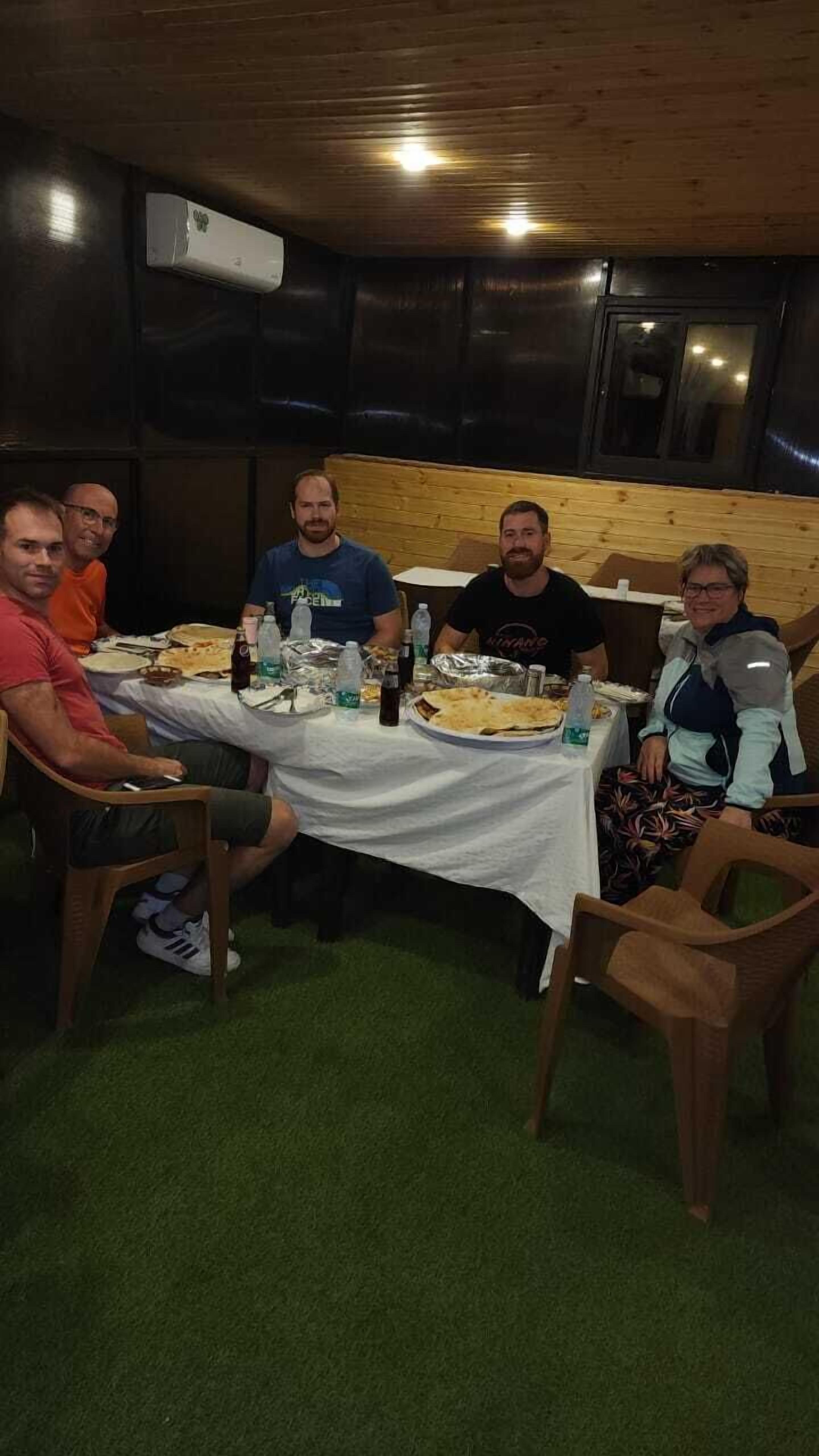 A group of people seated around a table with food and drinks, in a room with wooden walls and a ceiling fan.