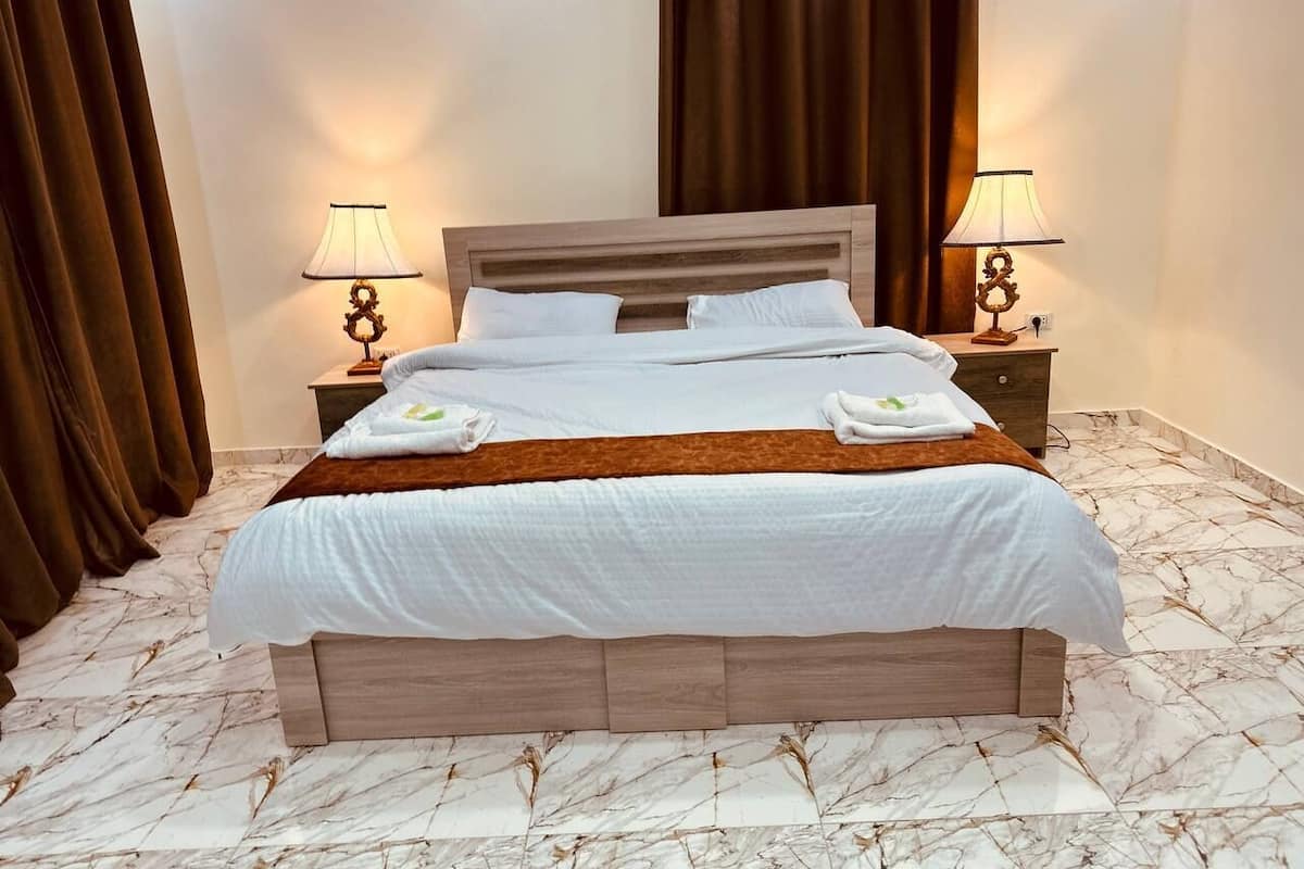 Una cama bien hecha con sábanas blancas y un plaid marrón, flanqueada por dos mesitas de noche con lámparas. La habitación tiene suelo de mármol y cortinas oscuras.