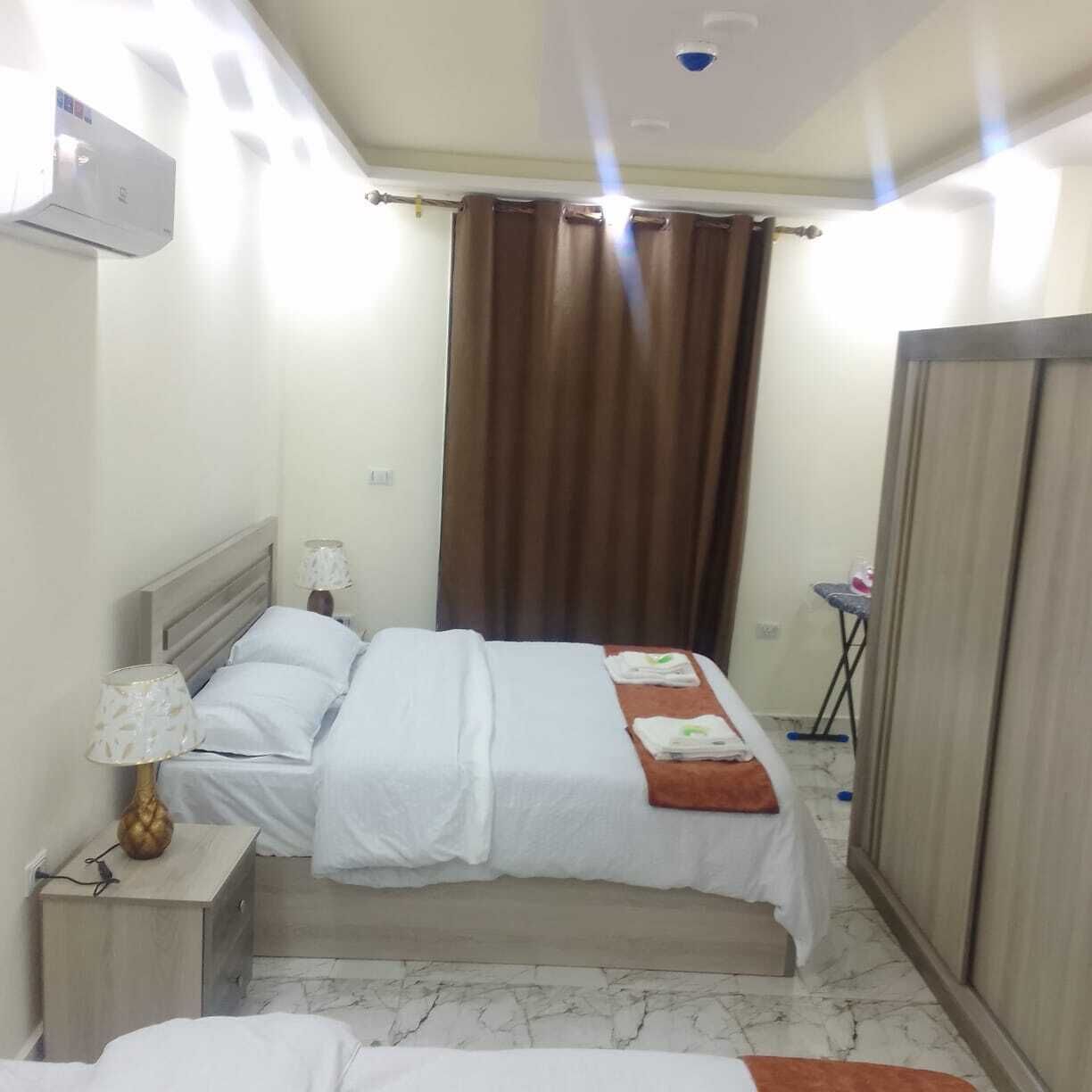 Quarto de hotel com cama, mesa de cabeceira com abajur, cortina e guarda-roupa.