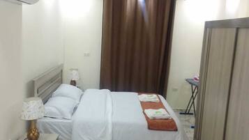 Quarto de hotel com cama, mesa de cabeceira com abajur, cortina e guarda-roupa.