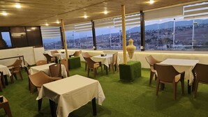 Ein Restaurant mit Tischen und Stühlen, einem grünen Teppich und großen Fenstern mit Blick auf eine Stadtlandschaft.
