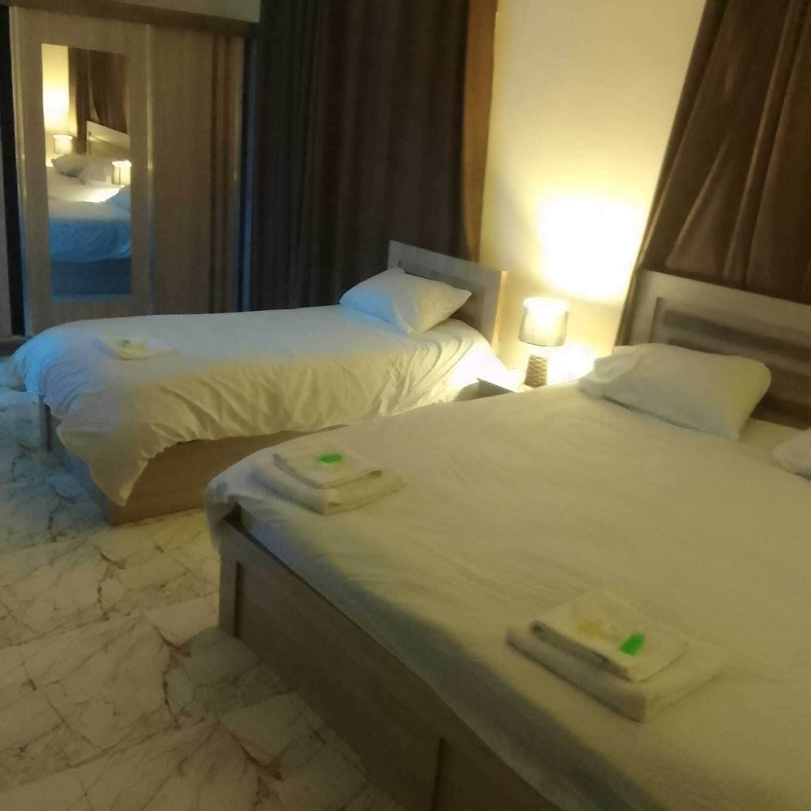 Dos camas ordenadas con sábanas y toallas blancas, una mesita de noche con lámpara y vistas a una habitación con ventana y cortinas.
