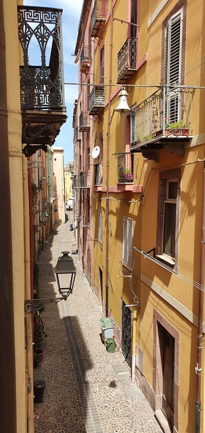 A narrow street with buildings on both sides, featuring balconies with railings and potted plants. - Single-family home with a large furnished patio; pets allowed (Bosa)