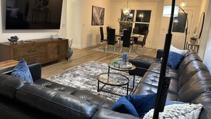 A modern living room with a leather sofa, a flat-screen TV, a dining area with a glass table, and a chandelier.