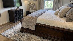 A bedroom with a large bed, a TV, a dresser, a chair, and a window with curtains.