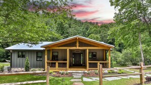 A cabin with a porch, surrounded by a stone pathway and a wooden fence, set in a lush green forest.