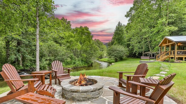 A fire pit surrounded by Adirondack chairs on a stone patio, overlooking a river and forested area with a cabin in the background.