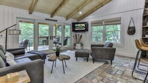 A spacious living room with a wooden ceiling, a television mounted on the wall, and a large window providing a view of the outdoors.