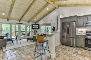 Una cocina amplia con un refrigerador y horno de acero inoxidable, una barra con taburetes, y vistas al exterior a través de ventanas grandes.