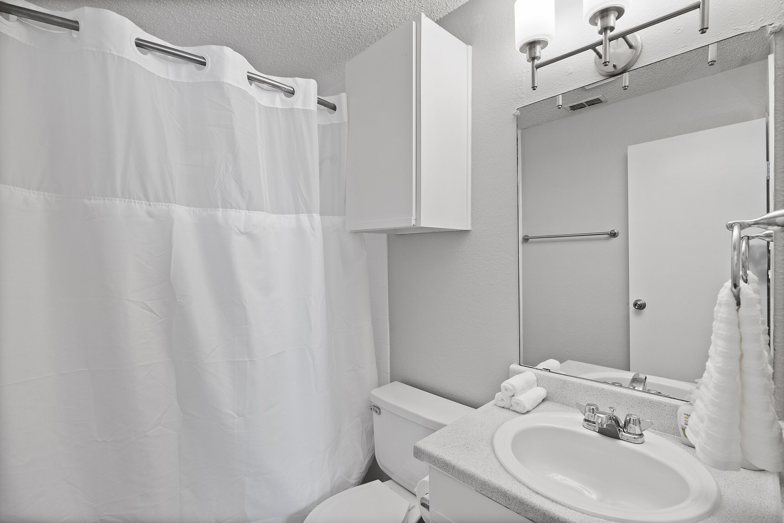 Un baño con cortina de ducha blanca, un inodoro, un lavabo con espejo y un perchero para toallas.