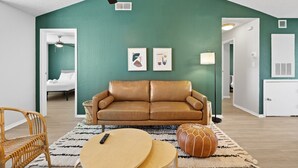 Ein Wohnzimmer mit einem braunen Ledersofa, einem runden Holztisch, einem Rattan-Sessel und einem gemusterten Teppich. An der Wand hängen zwei gerahmte Kunstwerke, eine Deckenlampe und im Hintergrund eine weiße Tür.