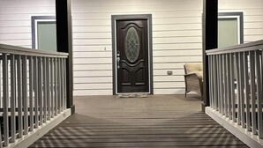 Eine Holzterrasse führt zu einer dunkel gefärbten Tür mit einer dekorativen Glasscheibe, gesäumt von weißen Geländern und einem Korbstuhl.