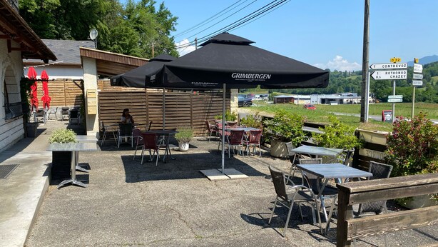 Un espace extérieur aménagé avec des tables et des chaises, protégé par un grand parasol, entouré d’une clôture en bois et agrémenté d’un panneau indiquant les directions vers les lieux d’intérêt à proximité.