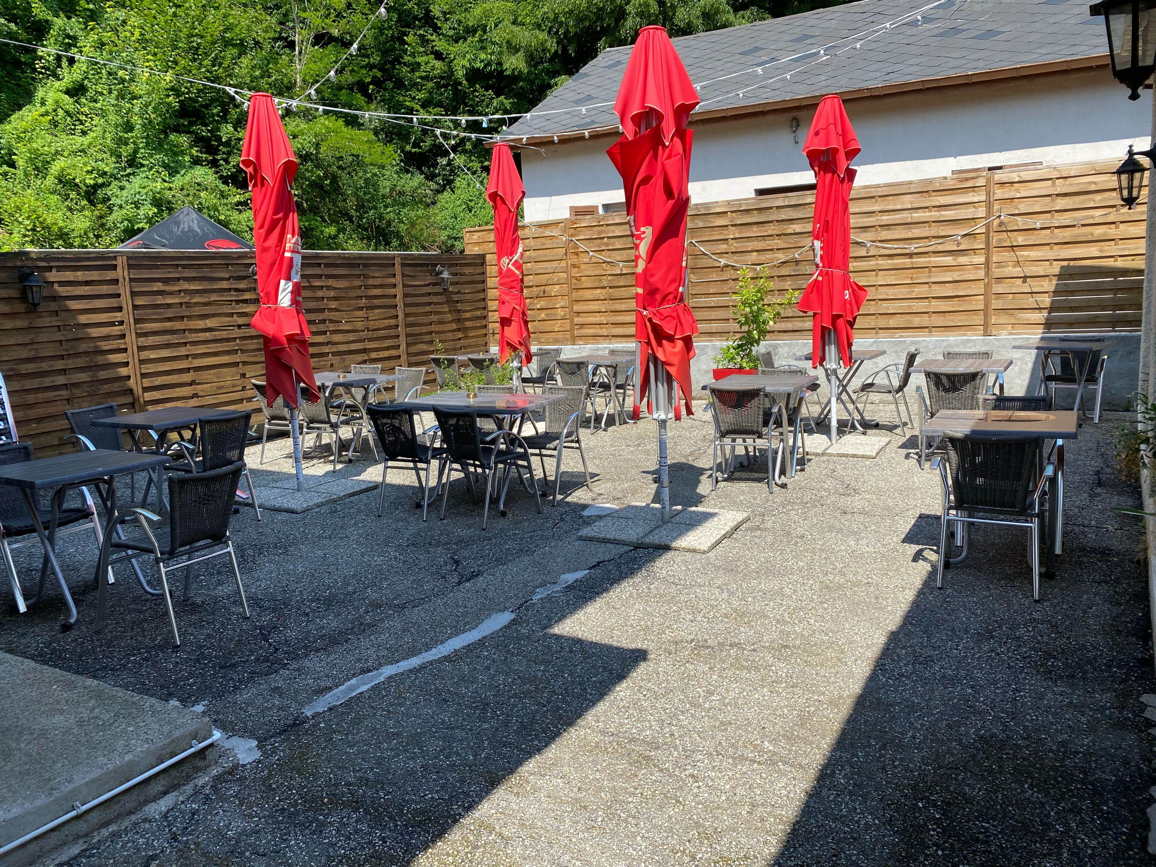 Un espace extérieur aménagé en terrasse, avec des tables et des chaises, des parasols rouges et une clôture en bois en arrière-plan.
