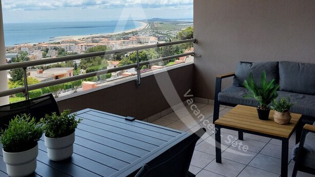 Un balcón con vista al mar y edificios, una mesa con plantas en macetas y un sofá gris.