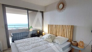 Un dormitorio con cama, mesita de noche, lámpara, una pequeña mesa y vistas al océano a través de una puerta corredera.