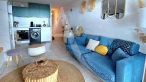 Una sala de estar moderna con un sofá azul, una mesa de centro redonda de madera y una cocina al fondo.