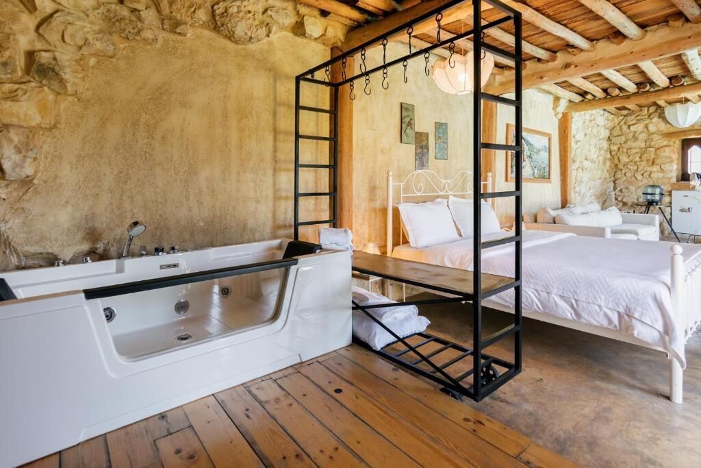 Un dormitorio rústico con pared de piedra, techo de madera, cama blanca, suelo de madera y bañera blanca.