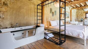 Un dormitorio rústico con pared de piedra, techo de madera, cama blanca, suelo de madera y bañera blanca.