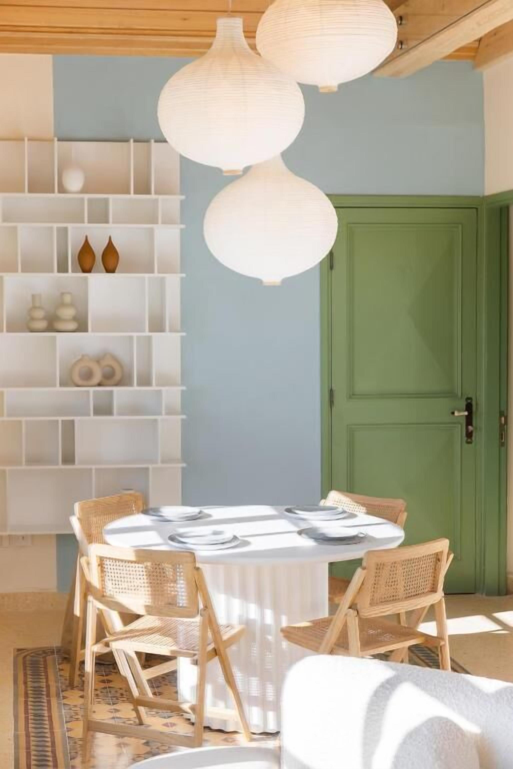 A dining area with a round table, wicker chairs, a white shelf with decorative items, and a green door.