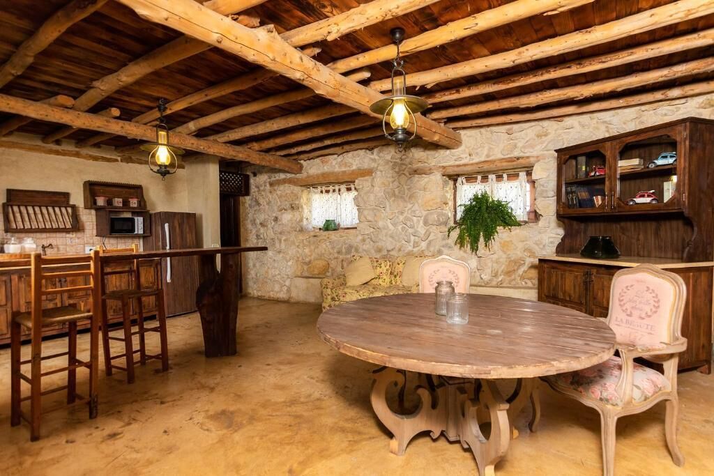 Un coin repas rustique avec un mur en pierre, des poutres en bois au plafond, une table ronde en bois et des chaises d’époque.