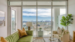 A modern living room with a green sofa, a coffee table, and a view of a cityscape through large windows. - @ Marbella Lane - Sleek Corner | Ocean View (Long Beach)