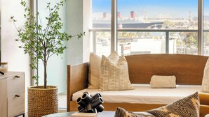 A modern hotel room with a large window, a sofa with patterned cushions, a small table with a plant and books, and a view of a cityscape. - @ Marbella Lane - Panoramic Delight Dream Escape! (Long Beach)