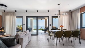 A modern living room with a sofa, dining table, and chairs, large windows with curtains, and a view of the sea. - Stylish property with heated 32sqm private pool, 5 bedrooms, 5 bathrooms, 1km from the beach (Podstrana)