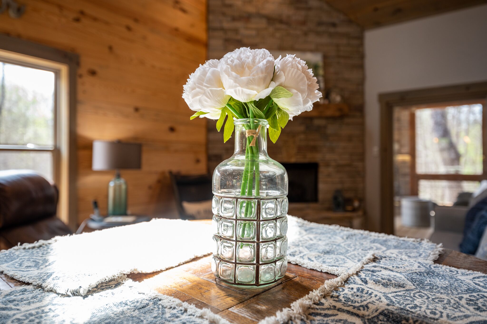 A clear glass vase with white flowers on a wooden table in a room with exposed brick walls and large windows.