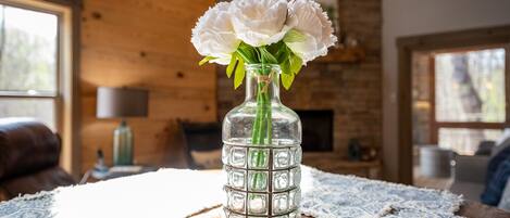 A clear glass vase with white flowers on a wooden table in a room with exposed brick walls and large windows.