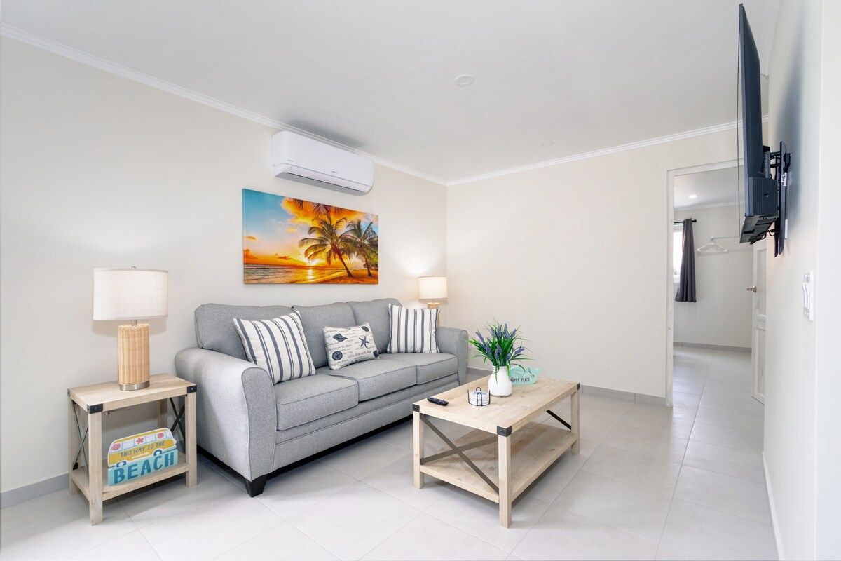 Uma sala de estar com um sofá cinza, duas mesas laterais, uma mesa de centro, uma pintura de um pôr do sol e uma TV de tela plana fixada na parede.