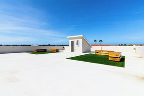 A modern building with a flat roof, a small entrance, and a patio area with wooden benches and artificial grass. - 2BR Condo w/ Shared Pool, near Palm Beach (Noord)