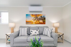A living room with a grey sofa, striped pillows, a wall-mounted painting of a sunset, and two side tables with lamps. - 2BR Condo w/ Shared Pool, near Palm Beach (Noord)
