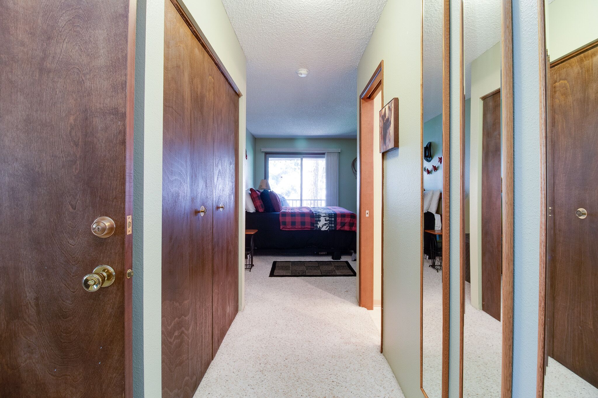 A narrow hallway leading to a bedroom with a bed, a window, and a door.