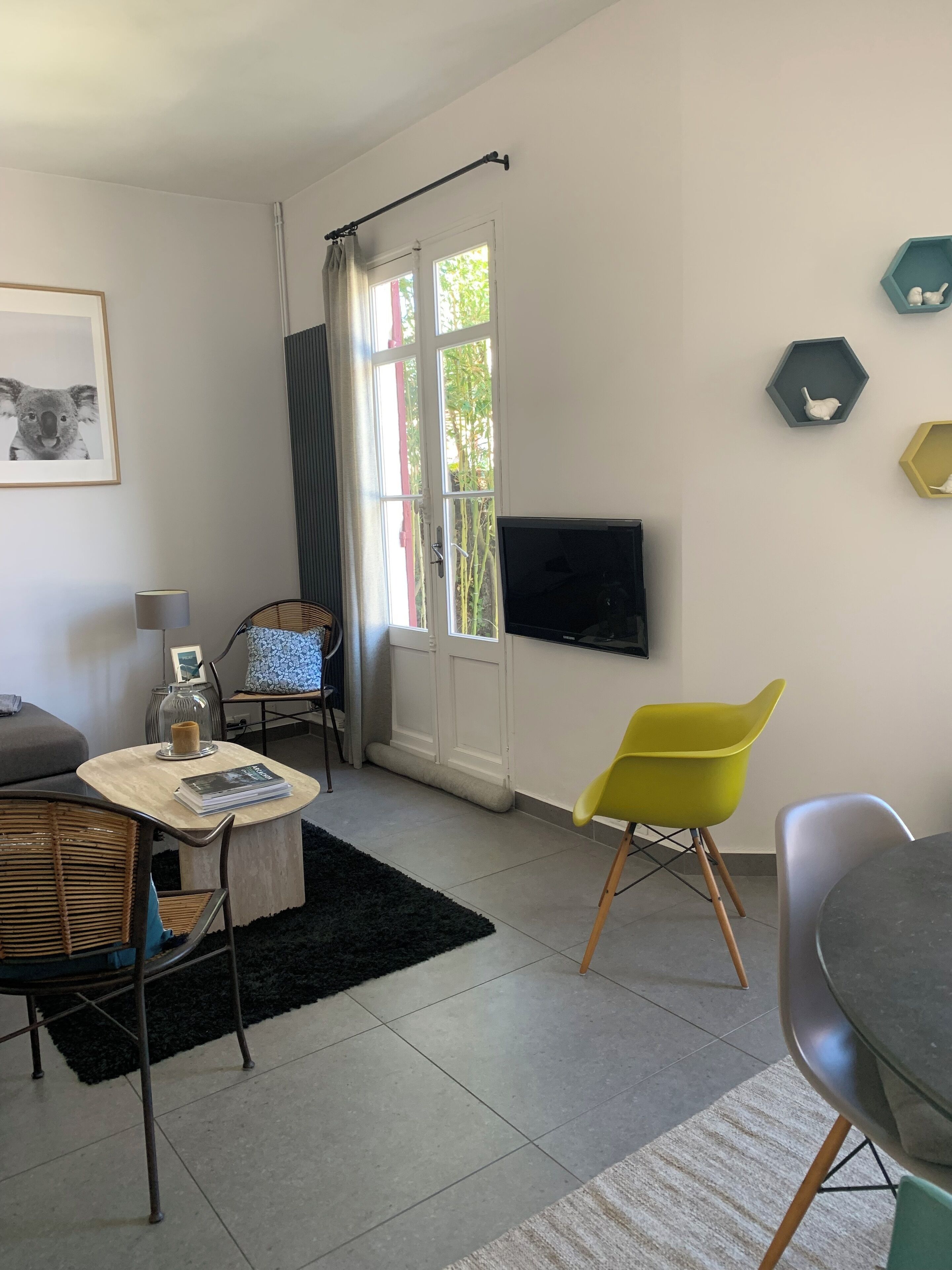 Ein modernes Wohnzimmer mit einer Couch, einem runden Tisch und einem gelben Sessel. Ein Fernseher ist an der Wand montiert und es gibt ein Fenster mit Vorhängen.