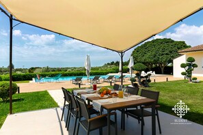 A covered outdoor dining area with a pool view, surrounded by greenery and a well-maintained lawn.
