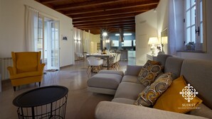 Un salón espacioso con techo de madera alto, un sillón amarillo, un sofá gris con cojines estampados y una zona de comedor al fondo.