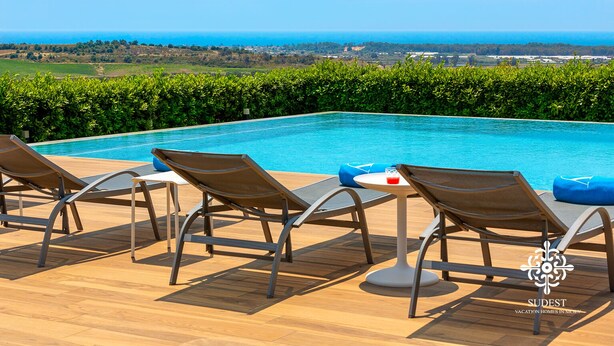 Zona junto a la piscina con sillas de descanso y una mesa, con vistas a un paisaje pintoresco y al mar.