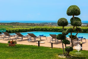 A poolside area with lounge chairs, a neatly trimmed hedge, and a view of the sea.