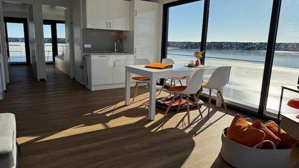 A modern kitchen with white cabinets and a dining area with a table and chairs. There is a large window with a view of the water. - Hausboot in Boltenhagen (Boltenhagen)